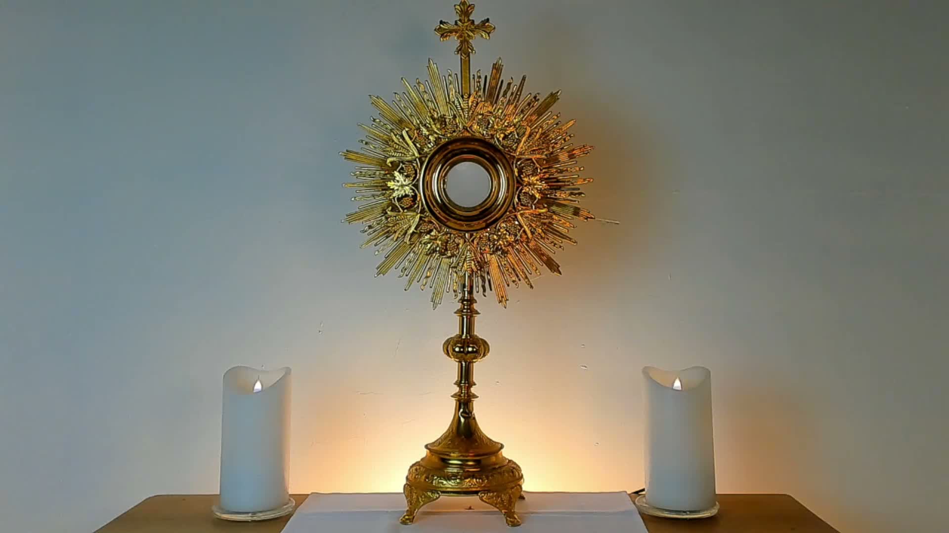 A golden monstrance featuring a sunburst design and a cross at its top stands centered on a wooden table with a white cloth, flanked by two lit white pillar candles, against a plain light-colored wall illuminated by a warm glow.