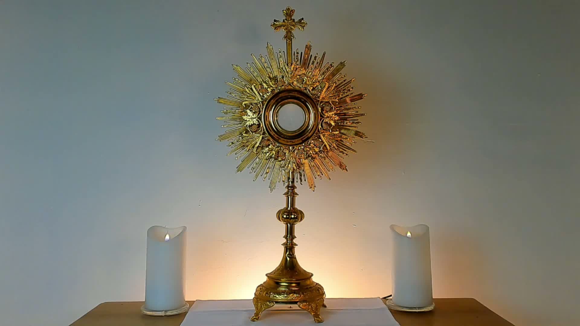 A golden, ornate monstrance with a radiant sunburst design and a cross on top stands centrally on a white cloth, flanked by two flickering white LED candles, all against a softly lit plain wall.