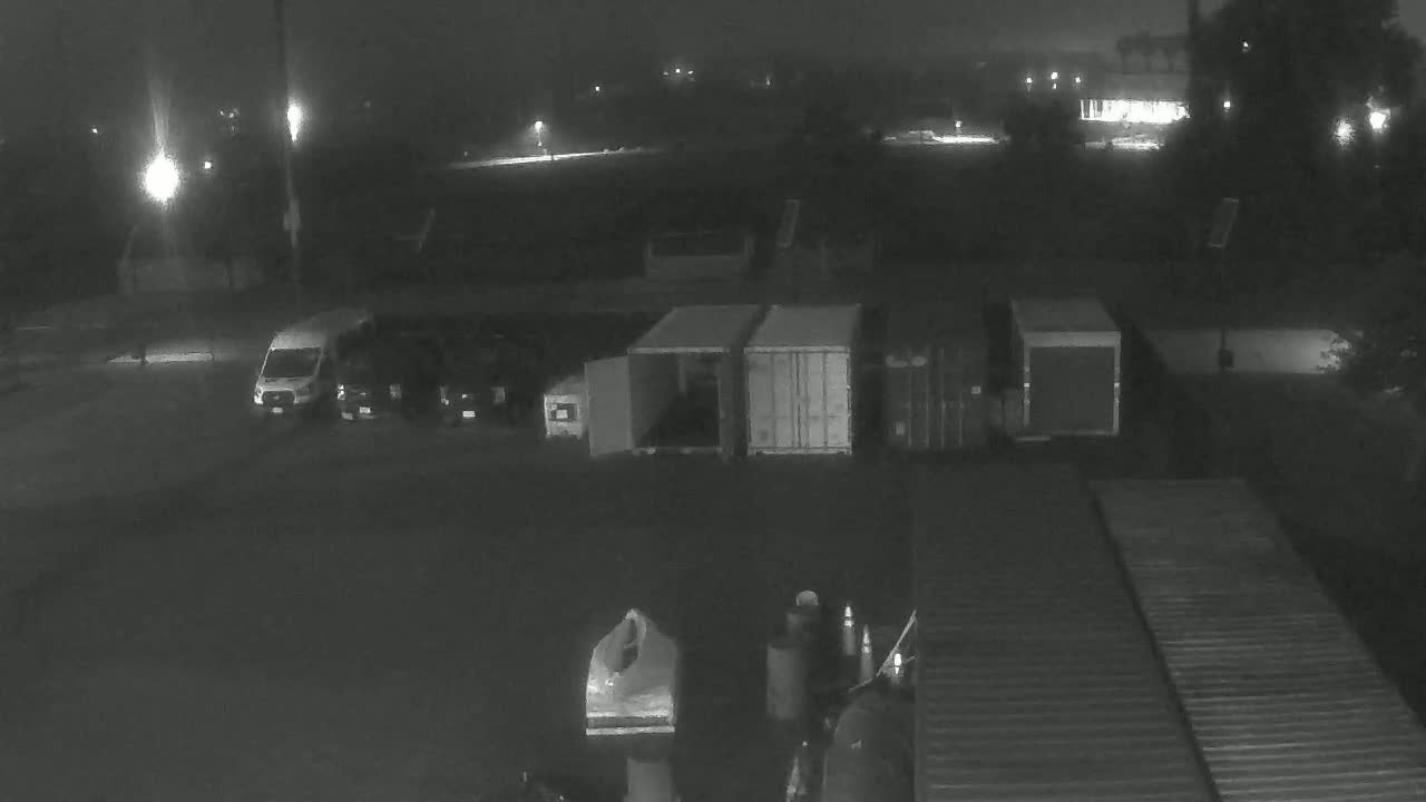 A nighttime, dimly lit parking lot with several vehicles, including a van and some cargo containers, under foggy or hazy conditions.