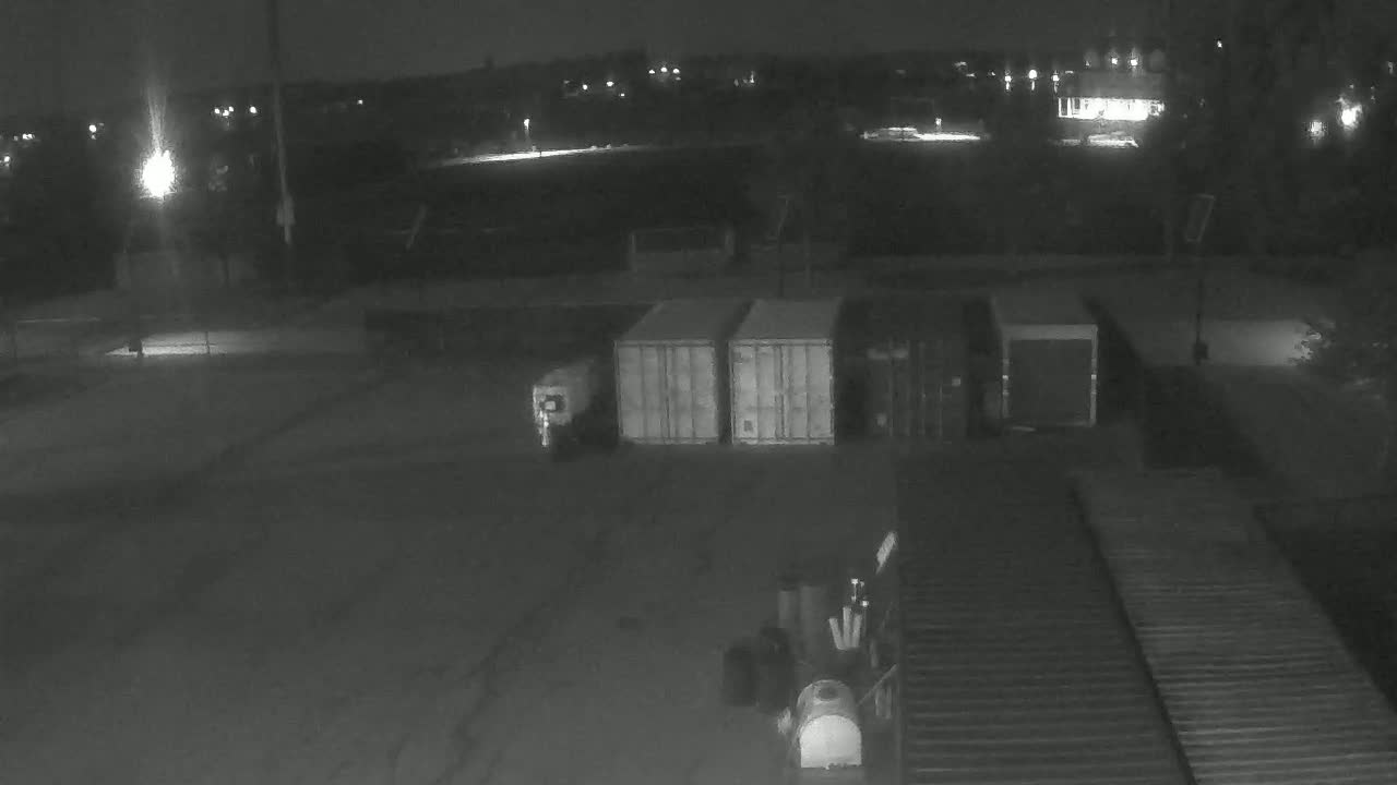 At night, a dark, empty lot contains several shipping containers, other equipment, and a vehicle, with a dimly lit background showing distant lights.