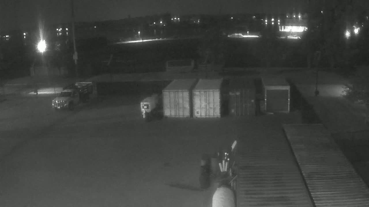 At night, a dark paved area contains several shipping containers, a truck, and a piece of machinery.