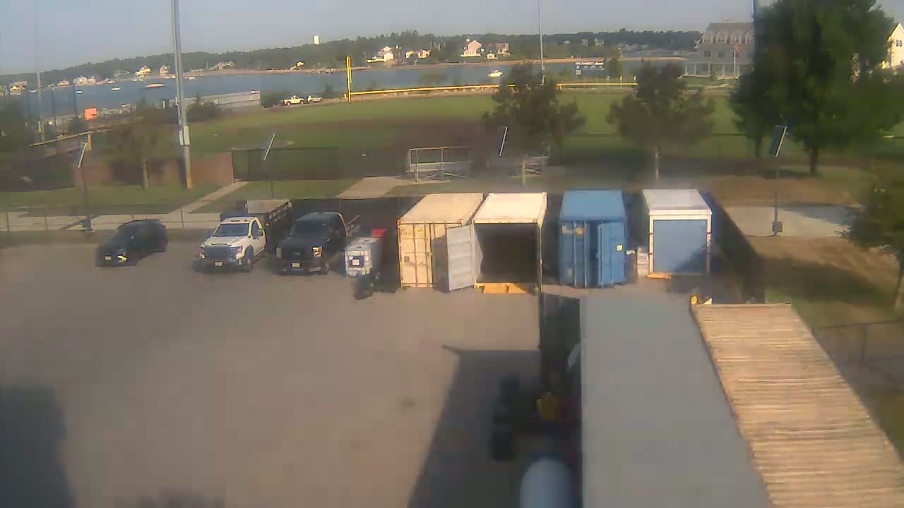 A view of a baseball field on an overcast day shows several shipping containers and two vehicles parked in a lot next to it.