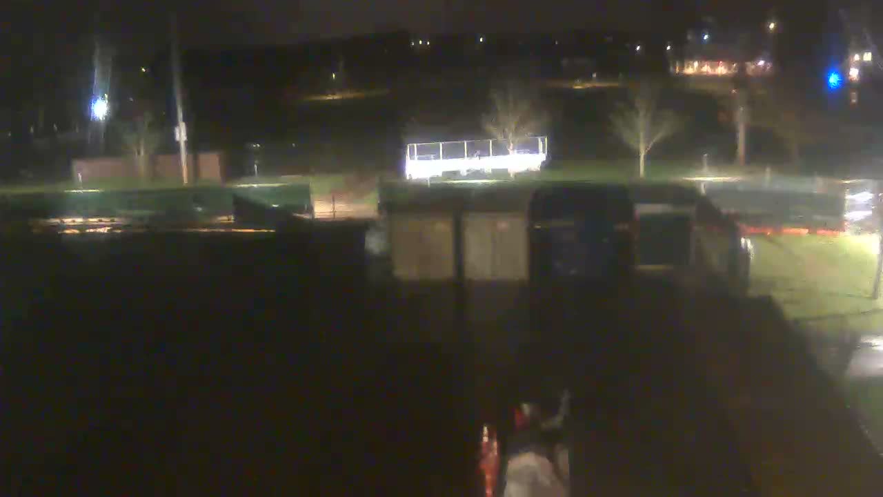 The blurry, low-light image depicts a damp night outdoors, showcasing a brightly lit white trailer behind two smaller, rectangular structures, with sparse trees and distant streetlights under a dark sky.