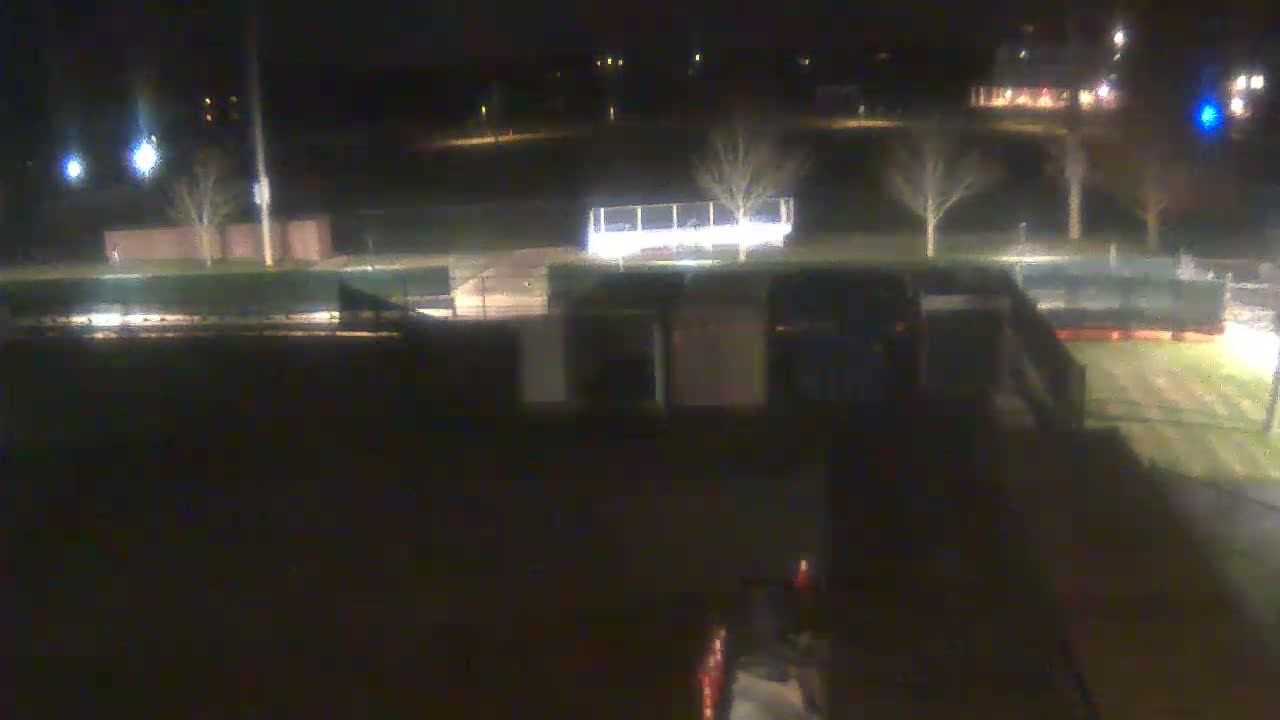 A blurry, dark night view shows an outdoor park or field featuring bare trees, various illuminated structures including bleachers, and distant artificial lights under a clear sky.