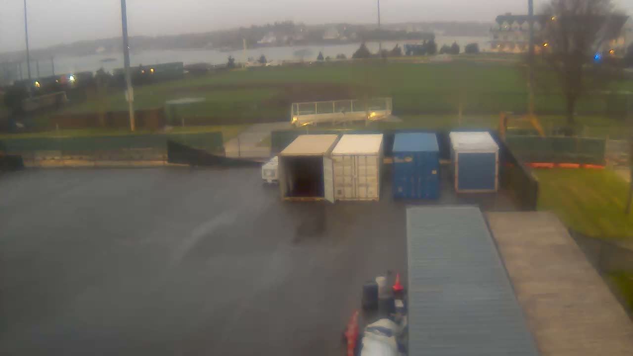 A blurry, dark night view shows an outdoor park or field featuring bare trees, various illuminated structures including bleachers, and distant artificial lights under a clear sky.