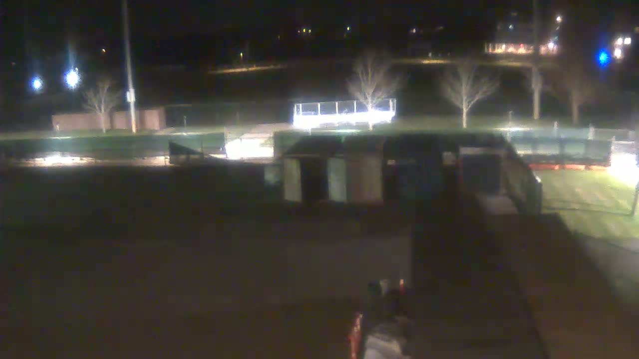 A dimly lit outdoor view at night shows a sports field or park with bare trees, dark ground, and a few small structures, under clear weather conditions.