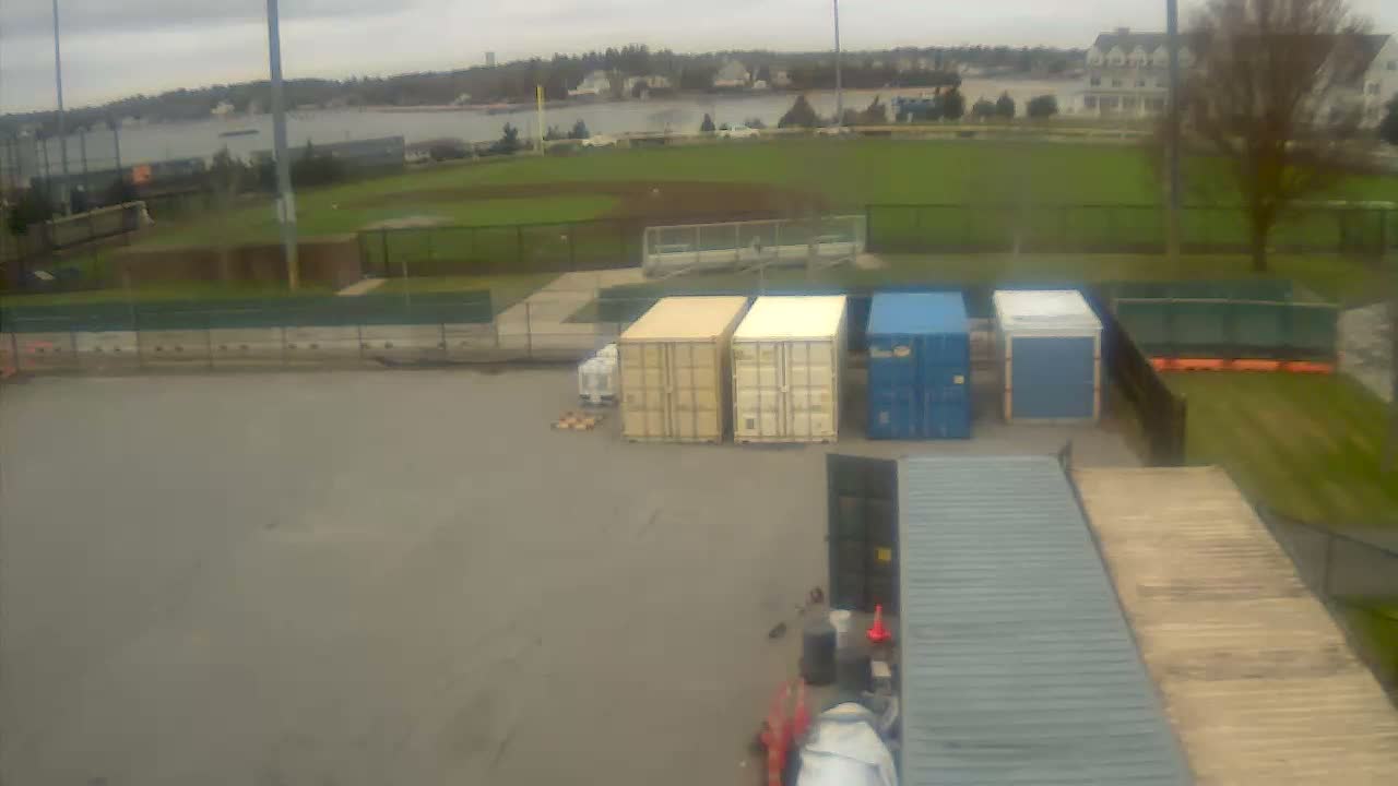 An outdoor scene features a paved area with multiple storage containers of various colors, adjacent to a green baseball field, with a body of water and distant buildings visible under an overcast sky.