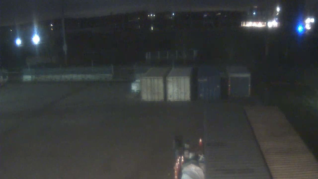 A dark and blurry night view of an outdoor industrial yard under clear skies reveals several shipping containers, a ribbed platform, and a piece of equipment with red lights in the foreground, all illuminated by scattered artificial light sources.