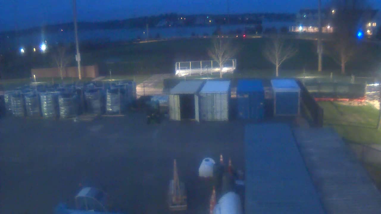 A dark and blurry night view of an outdoor industrial yard under clear skies reveals several shipping containers, a ribbed platform, and a piece of equipment with red lights in the foreground, all illuminated by scattered artificial light sources.