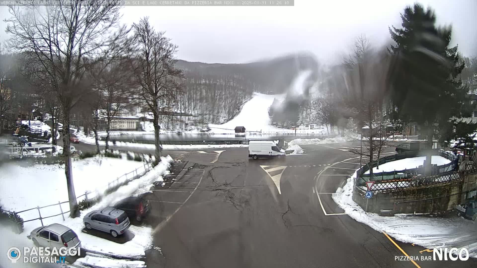 A snowy parking lot and roadway are seen with several cars and a van, next to a snow-covered wooded area with a ski slope visible in the background, under overcast skies.