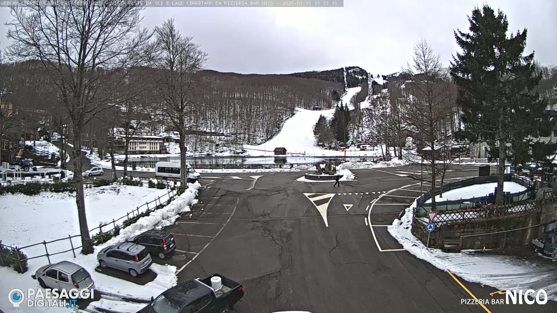 A snowy parking lot and roadway are shown with several cars parked, overlooking a snow-covered ski slope and frozen lake, under an overcast sky.