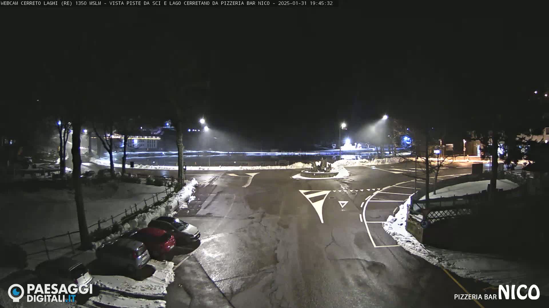 A nighttime view of a snow-covered parking lot and roadway intersection, illuminated by streetlights.
