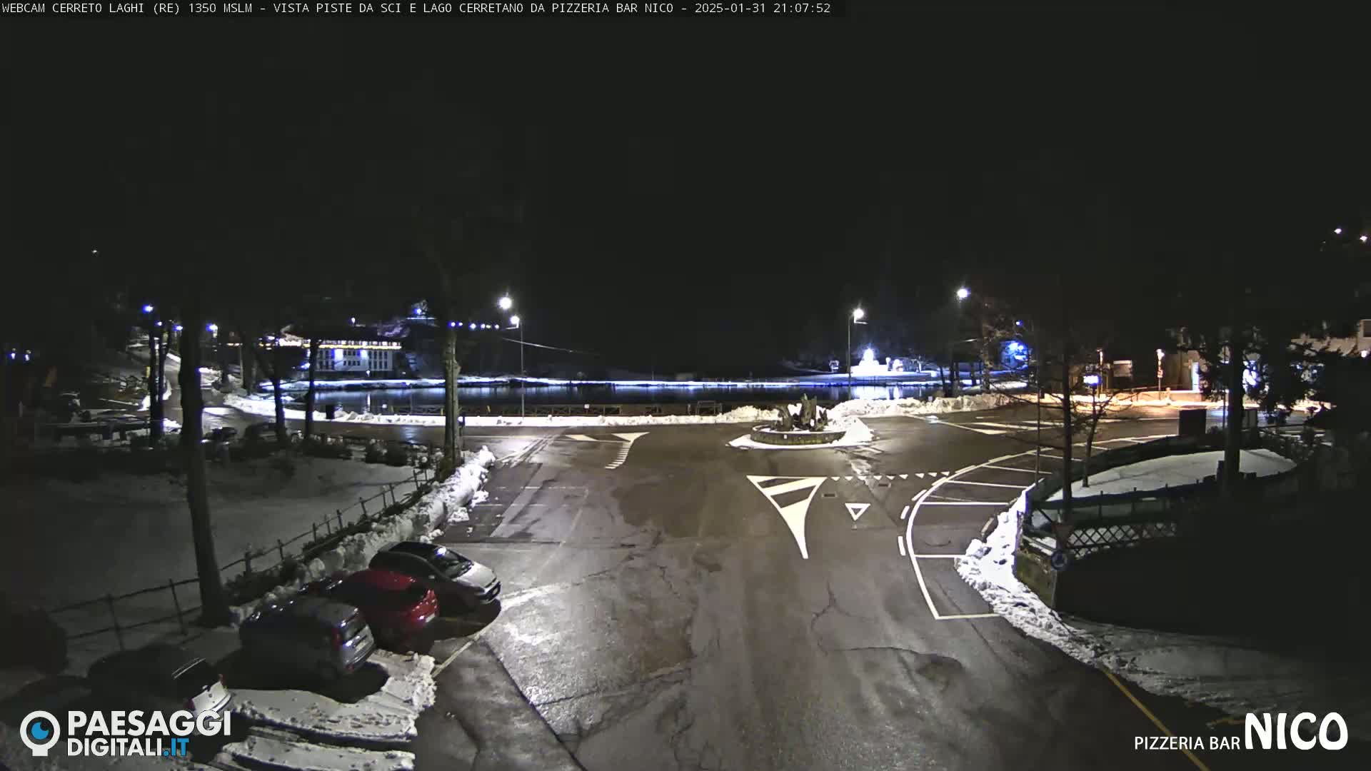 A nighttime view of a snow-dusted parking lot and roadway surrounding a small body of water, with several cars parked, and some buildings visible in the background under a clear, dark sky.