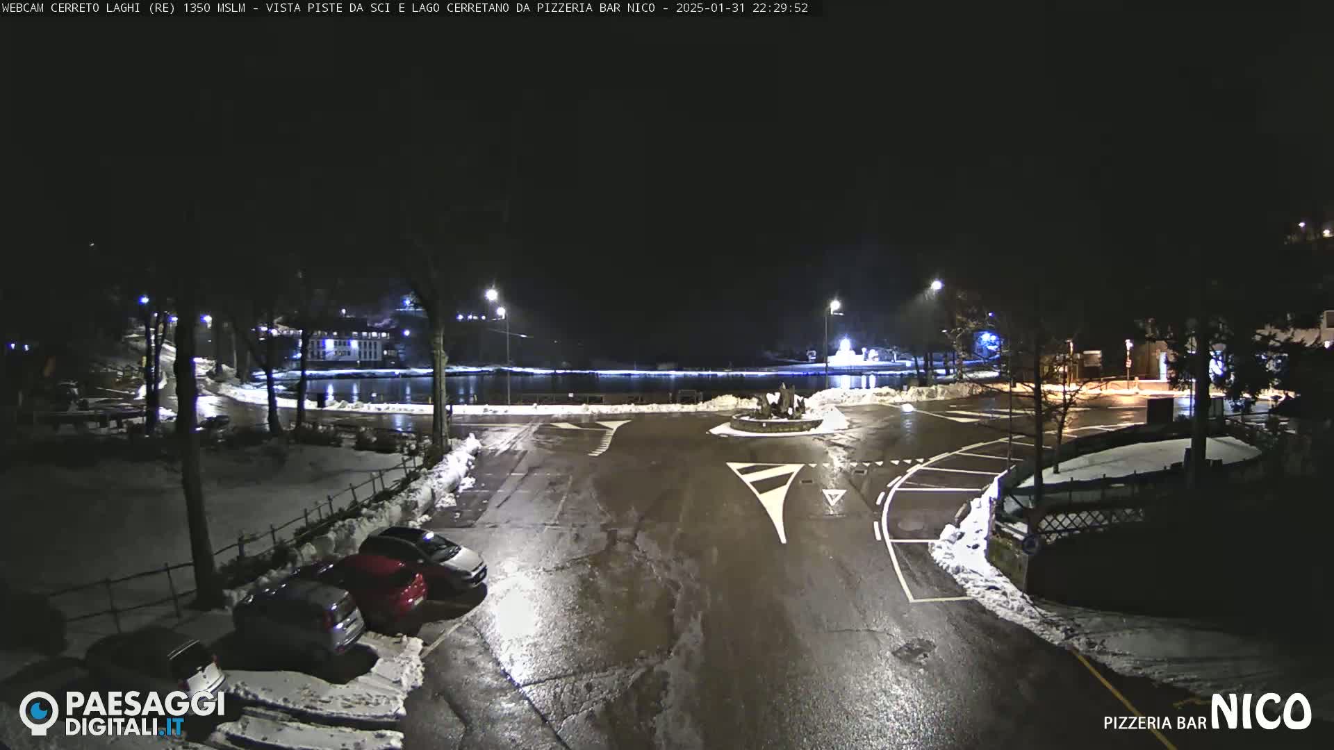A nighttime view of a parking lot and roundabout near a body of water, partially covered in snow, with several cars parked.