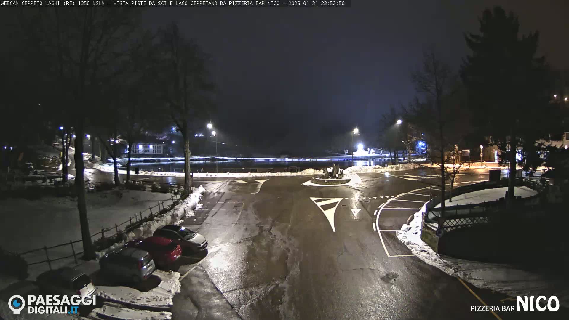 A nighttime view of a snow-dusted parking lot and roundabout near a body of water, with cars parked and streetlights illuminating the scene.