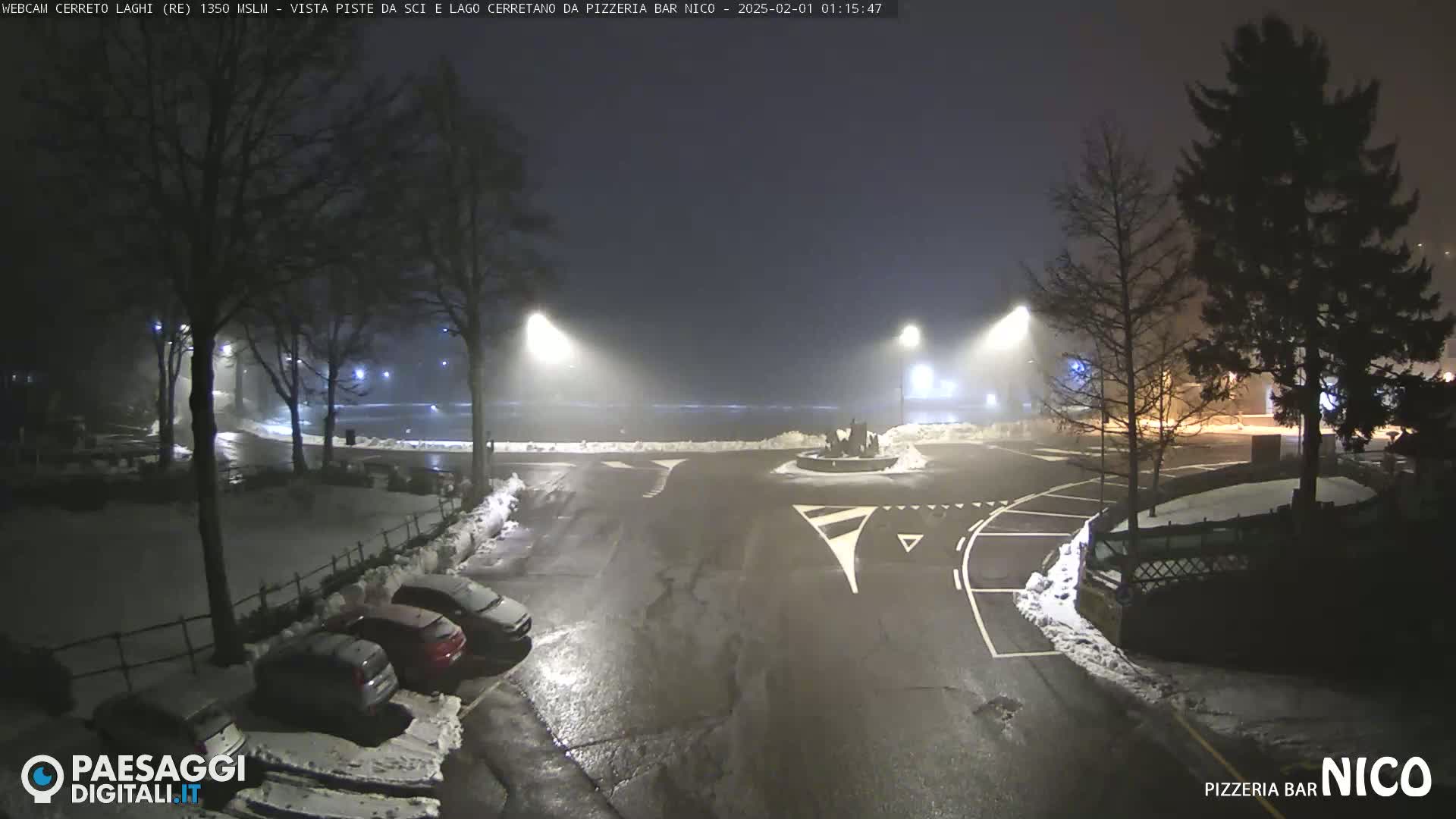 A nighttime parking lot with snow on the ground and fog in the distance, illuminated by streetlights.