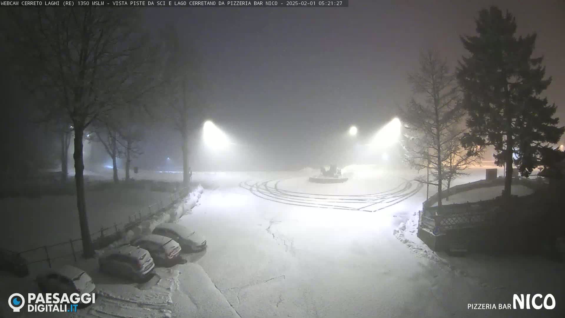 A snow-covered parking lot at night is dimly lit by streetlights in foggy conditions, with several cars parked and tire tracks visible in the snow.
