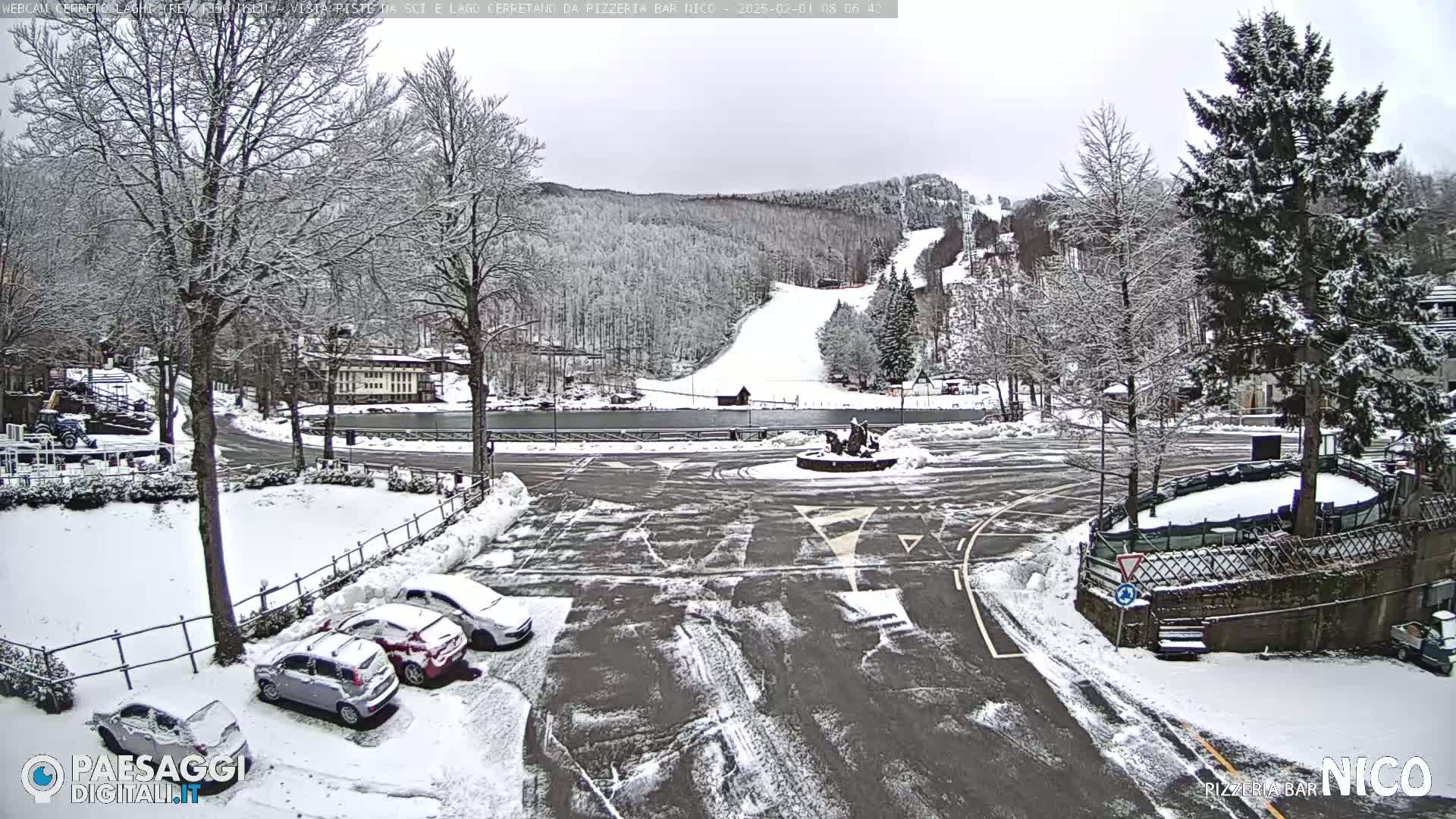 A snow-covered parking lot and roundabout are situated in front of a building, with a snowy ski slope and wooded hills visible in the background under an overcast sky.