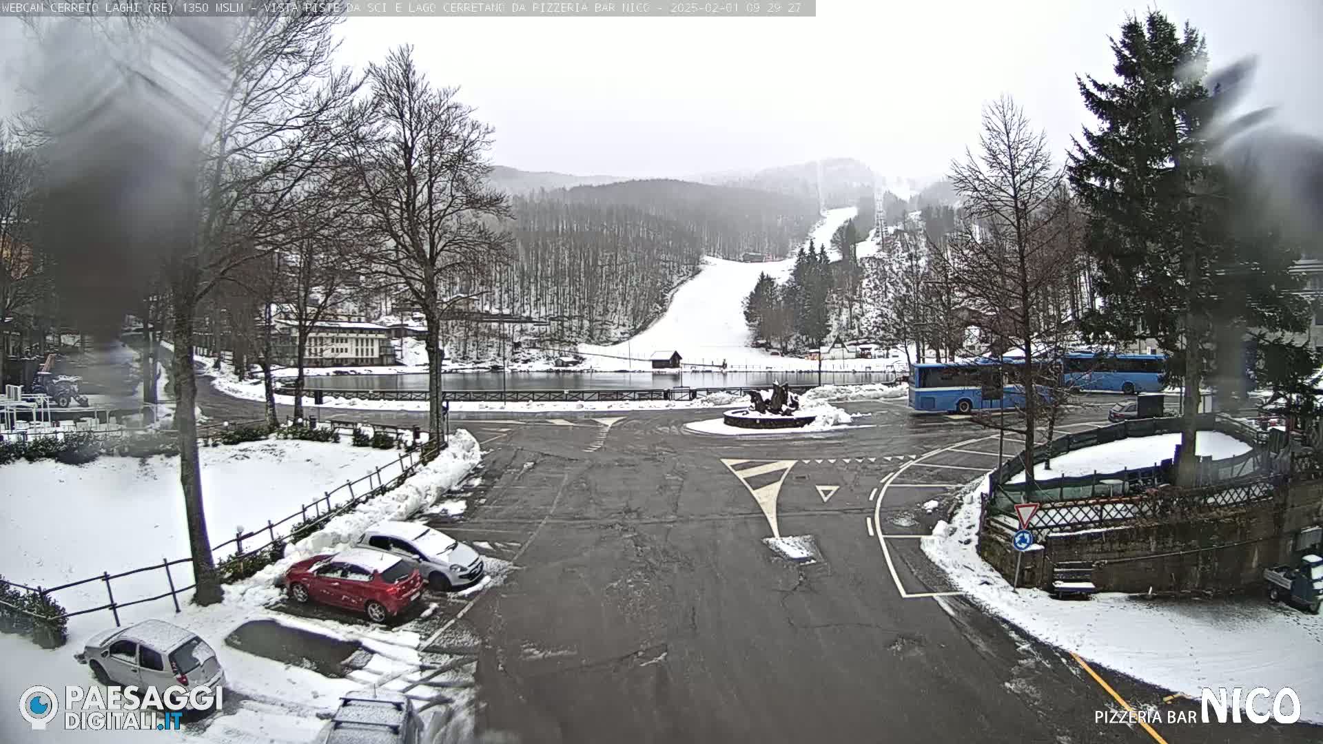 A snowy parking lot and roundabout with a few cars and a bus, overlooking a snow-covered ski slope and a frozen lake under an overcast sky.