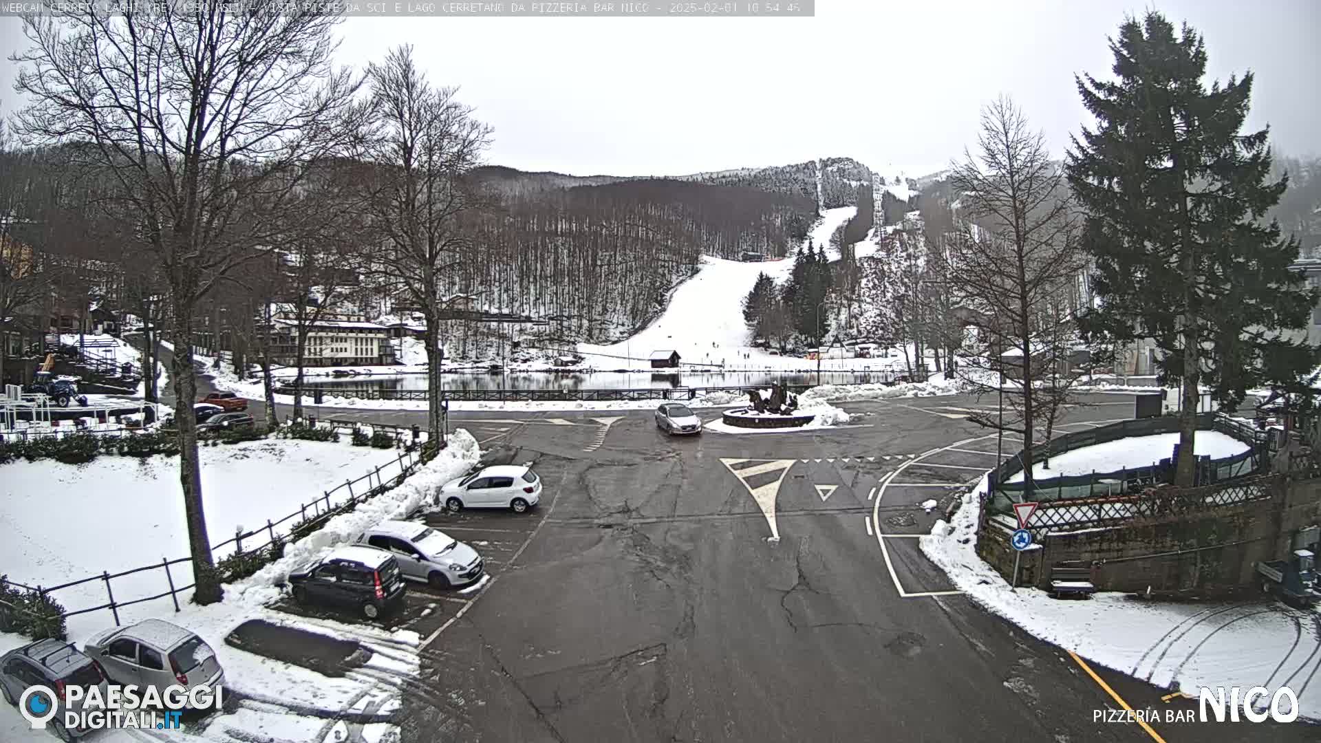 A snow-covered parking lot with several cars, surrounded by snow-dusted trees and buildings, overlooks a ski slope on a cloudy day.