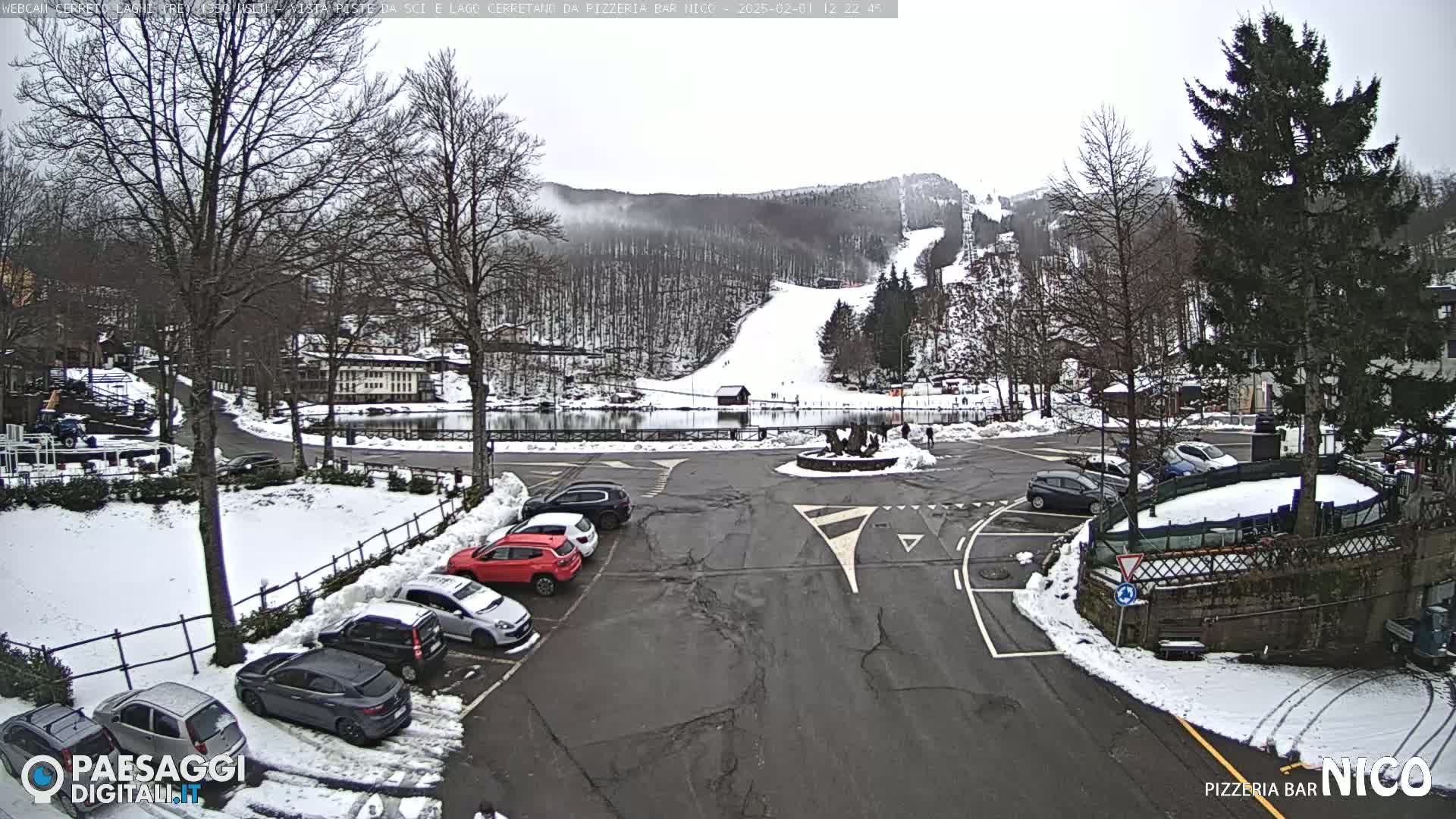 A snow-covered parking lot with several cars is situated near a small lake and a ski slope on an overcast day.