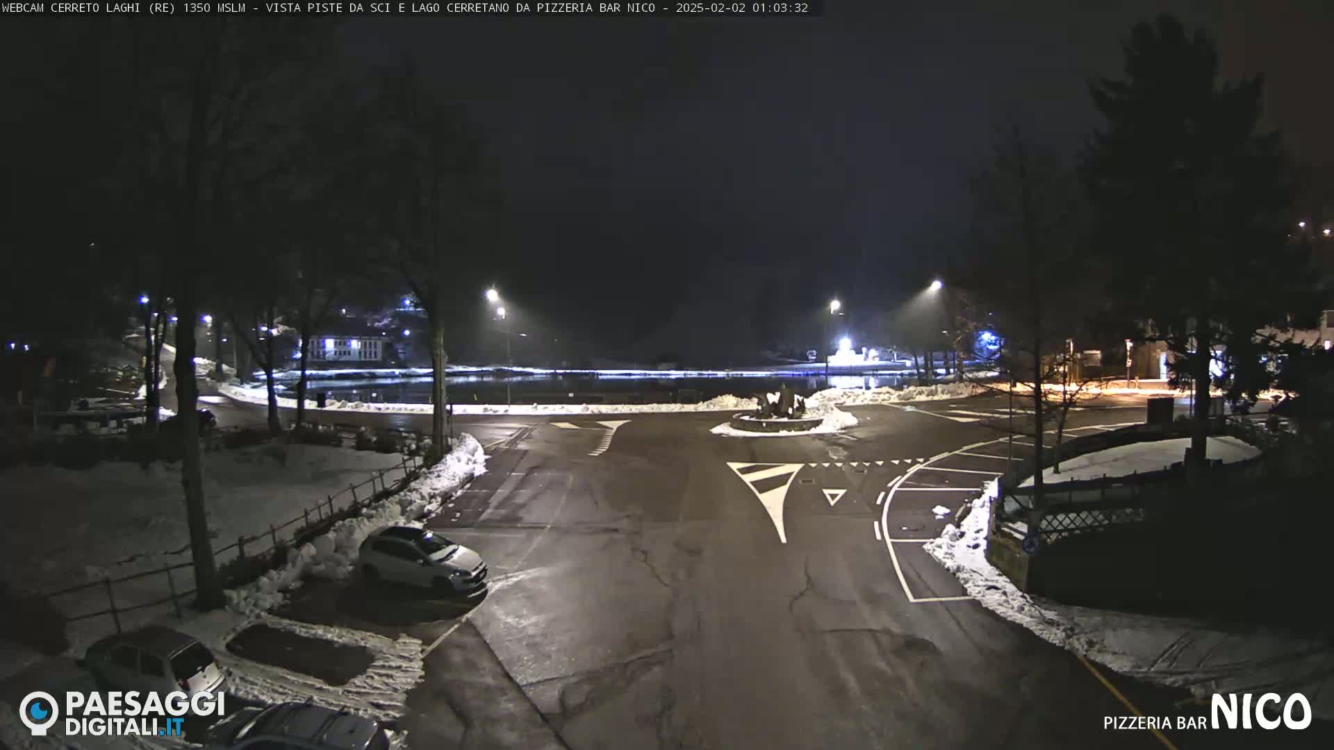 A nighttime view of a snow-covered parking lot and roundabout near a body of water, with cars parked and streetlights illuminating the scene.