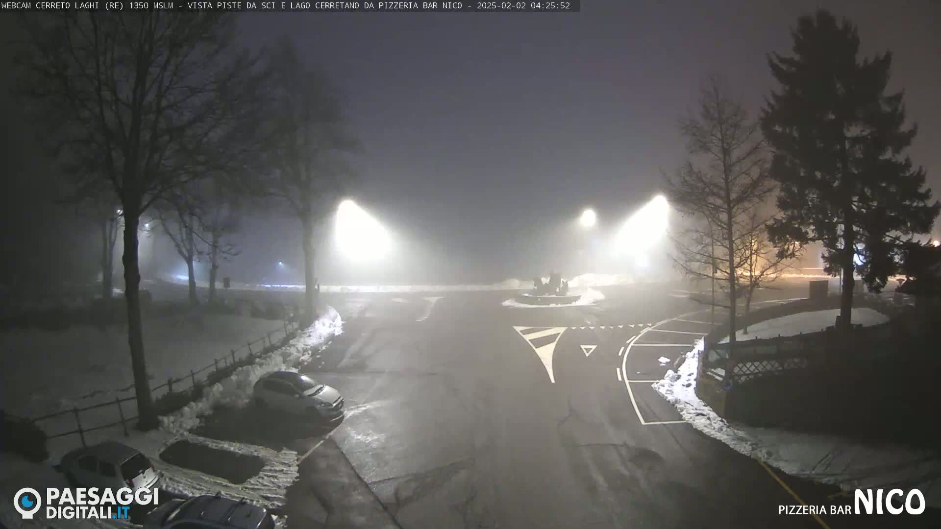 A foggy night scene shows a parking lot with a few cars and snow on the ground, illuminated by streetlights.