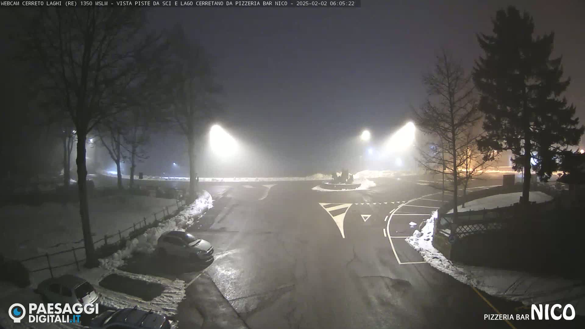 A foggy, nighttime scene shows a parking lot and roadway with a few cars, illuminated by streetlights, with snow on the ground and some trees.