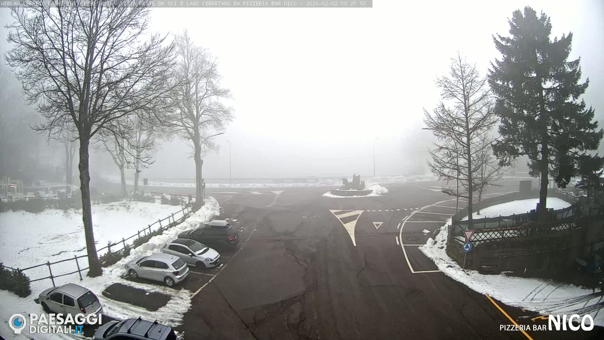 A foggy parking lot with several cars parked in it, surrounded by bare trees and patches of snow.