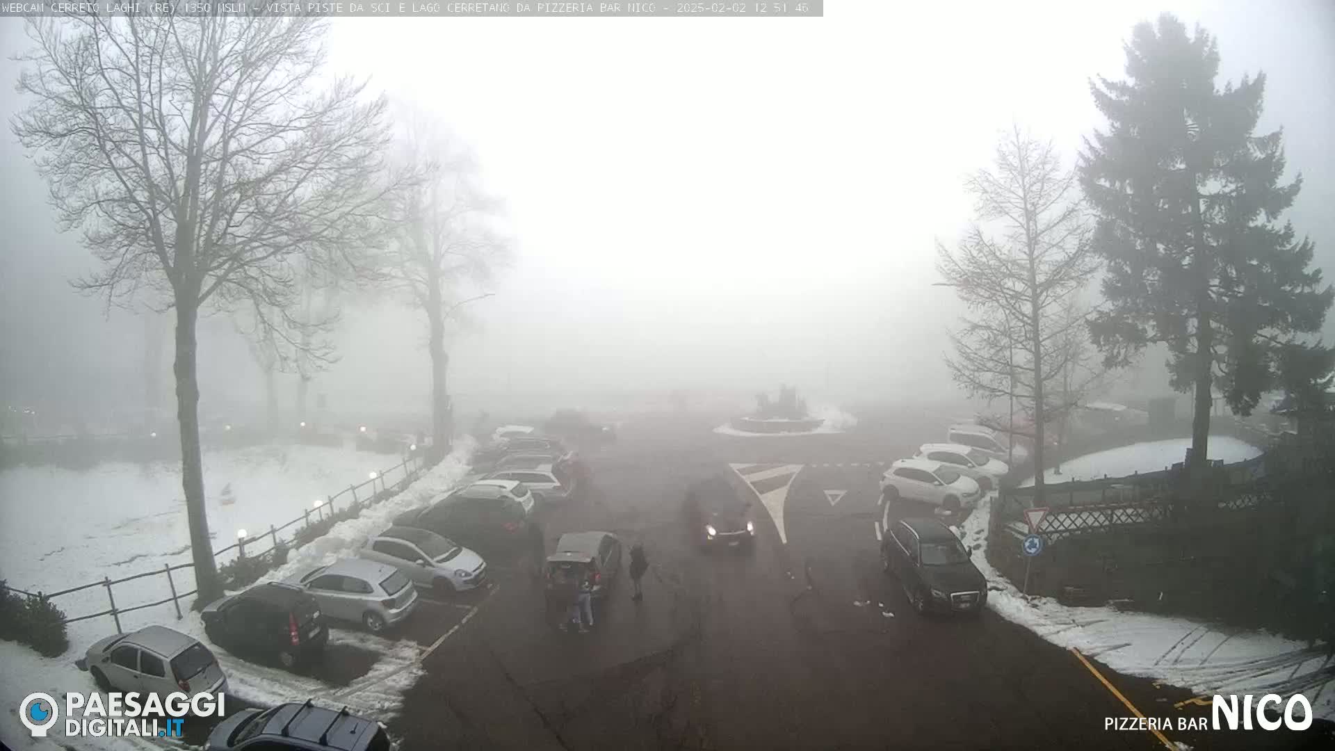 A foggy parking lot with cars and a few people, surrounded by snow-covered ground and bare trees.
