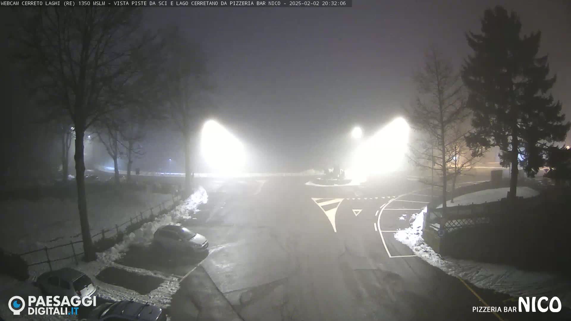 A foggy night scene shows a snow-covered parking lot and roadway illuminated by bright lights, with several cars parked and trees visible in the background.