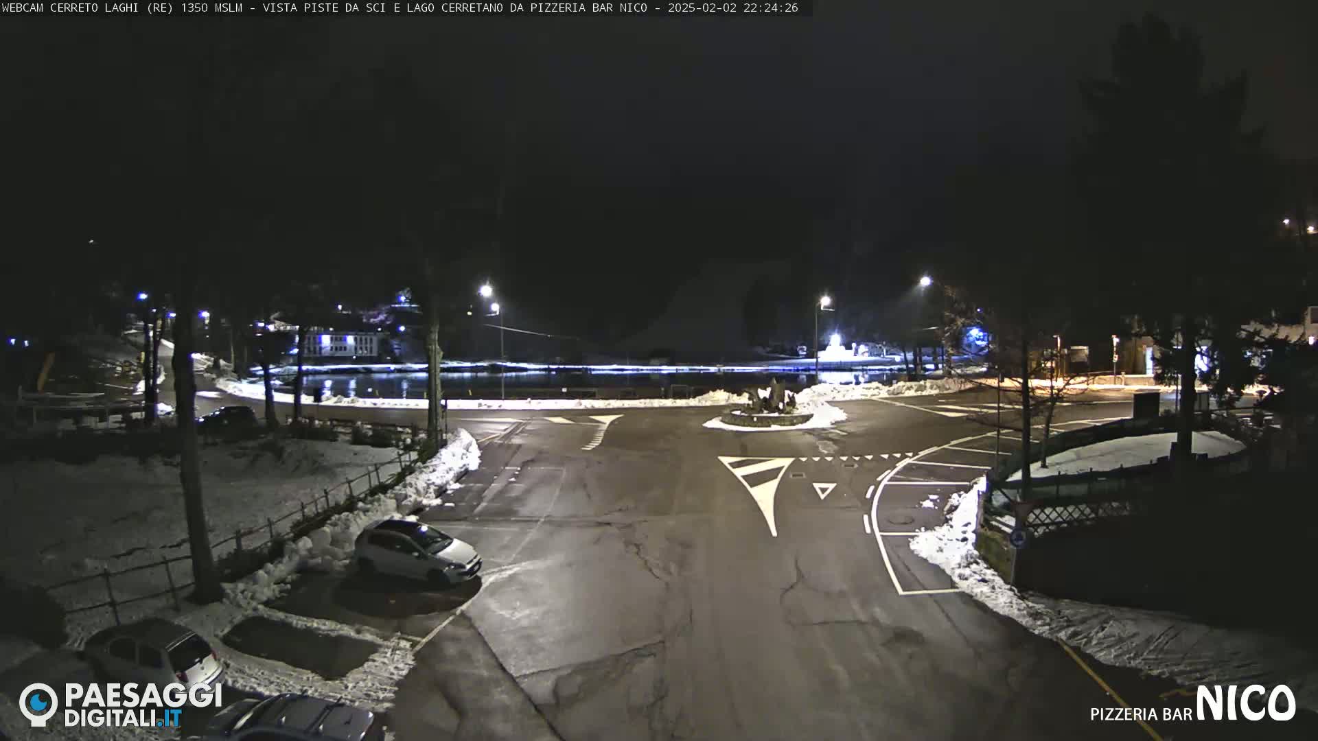 A nighttime, snow-covered parking lot and roadway surrounding a small roundabout near a body of water, illuminated by streetlights.