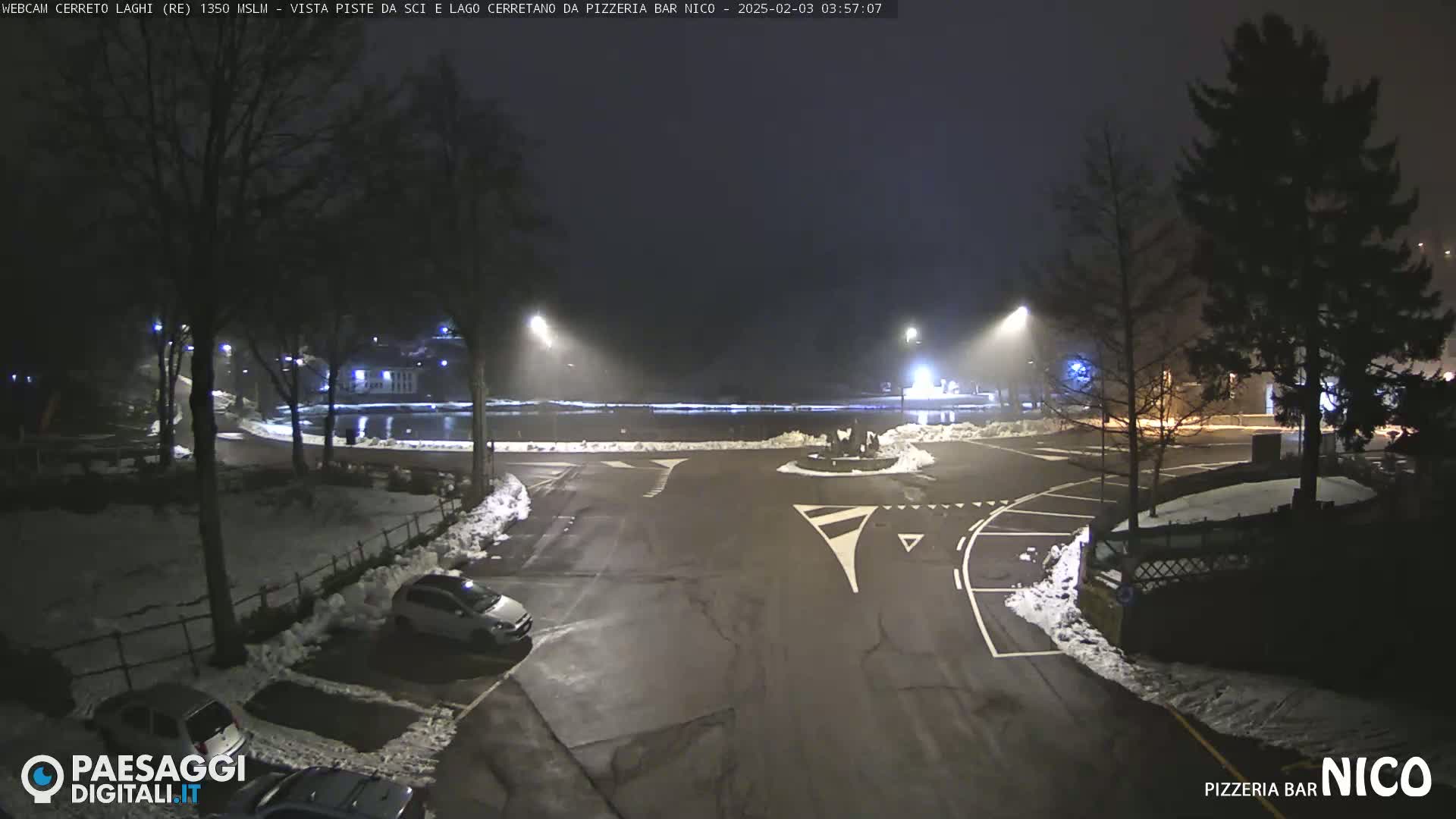 A nighttime view of a snow-dusted parking lot and roadway near a body of water, under foggy conditions, with several cars parked and streetlights illuminating the scene.