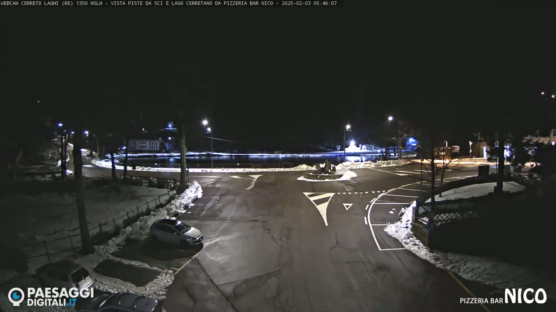 A nighttime view of a parking lot and roadway intersection with some snow on the ground and parked cars.