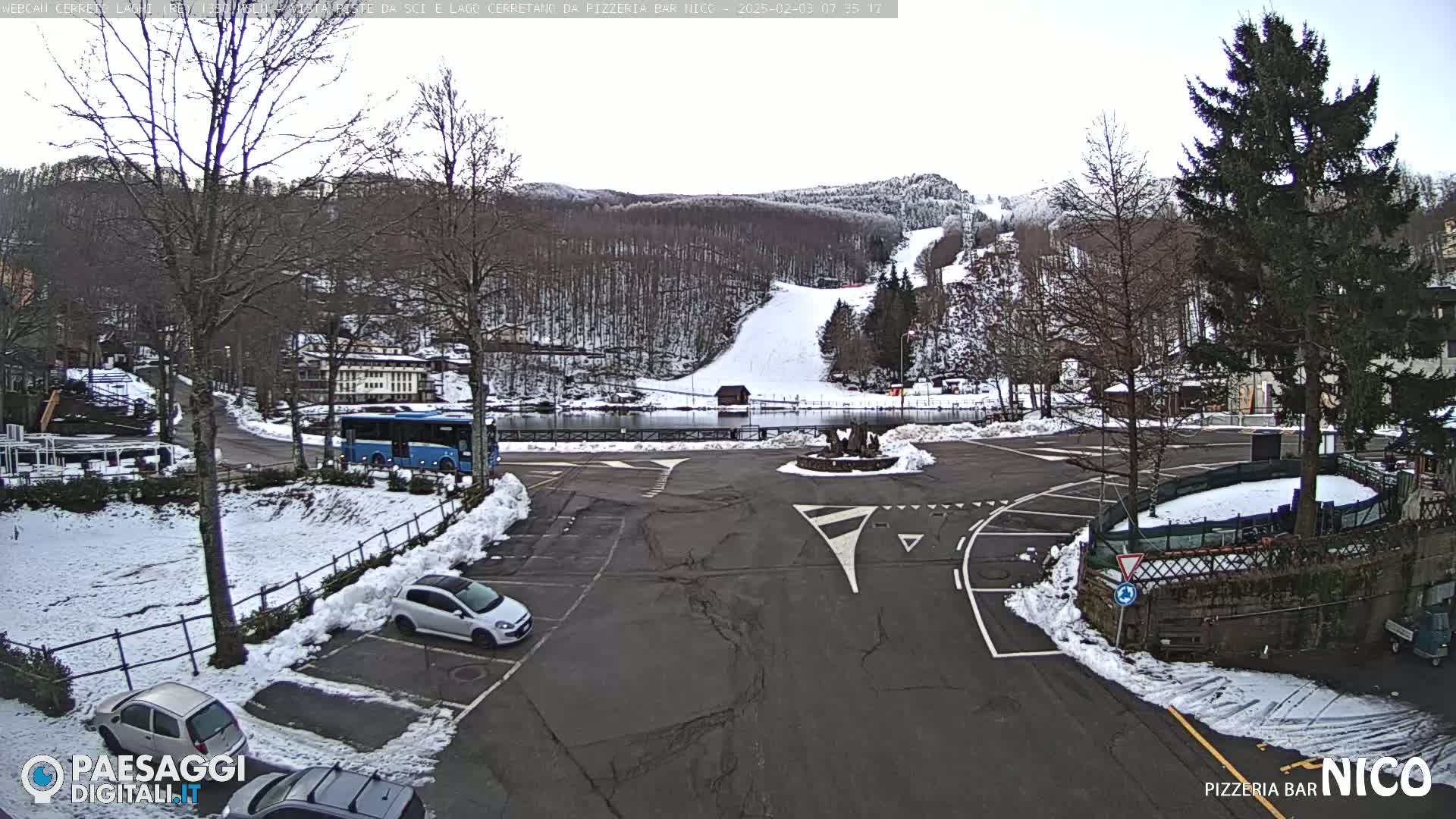 A snow-covered parking lot and roadway are situated below a snowy hillside with a ski slope, and a bus is parked nearby.