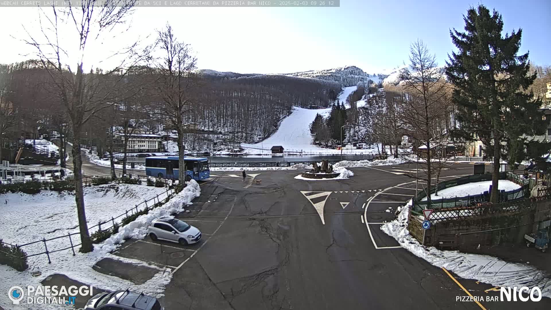 A parking lot with a few cars and a bus, next to a snow-covered road leading to a ski slope on a sunny day.