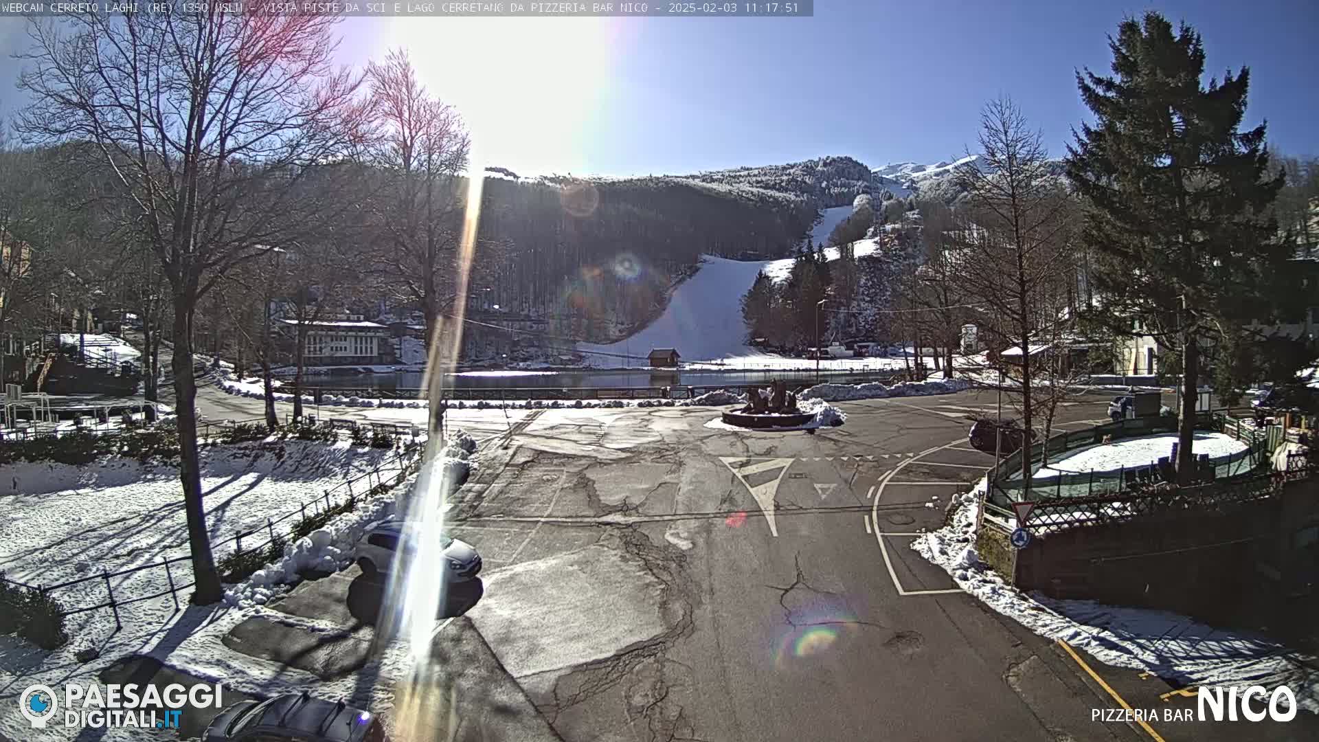 A sunny, snow-dusted parking lot overlooks a ski slope and a lake nestled in a mountainous landscape.