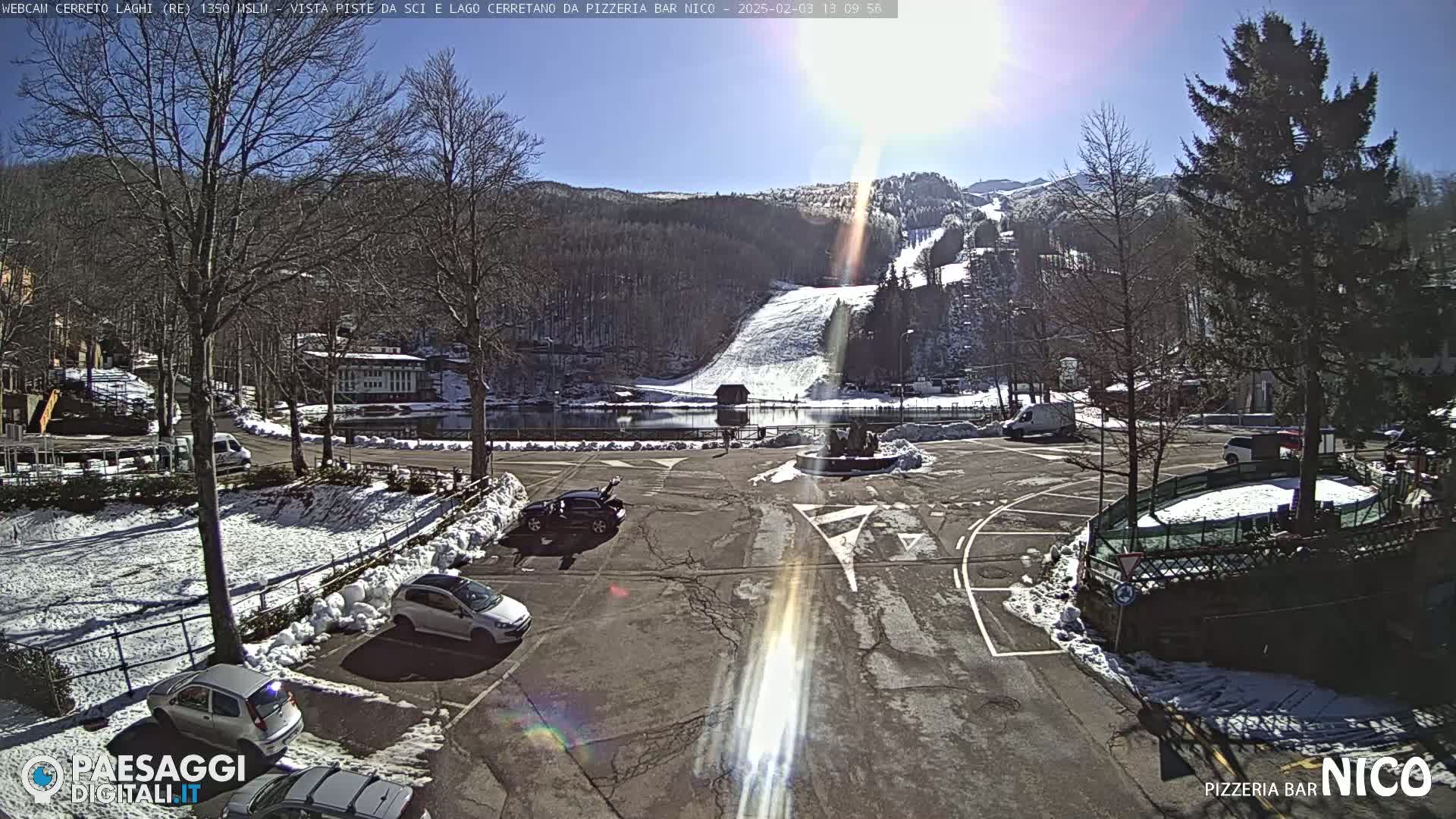 A sunny winter day shows a parking lot with several cars, some snow on the ground, and a view of ski slopes and a lake beyond.