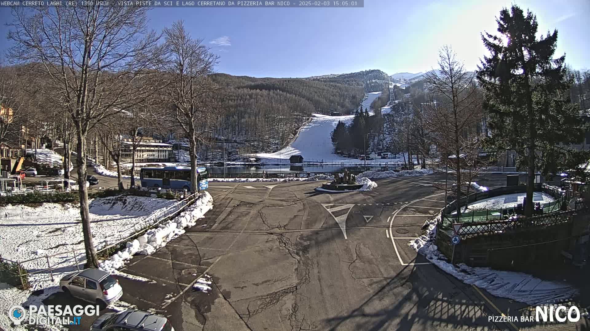 A sunny parking lot with snow on the ground and nearby ski slopes visible in the background.