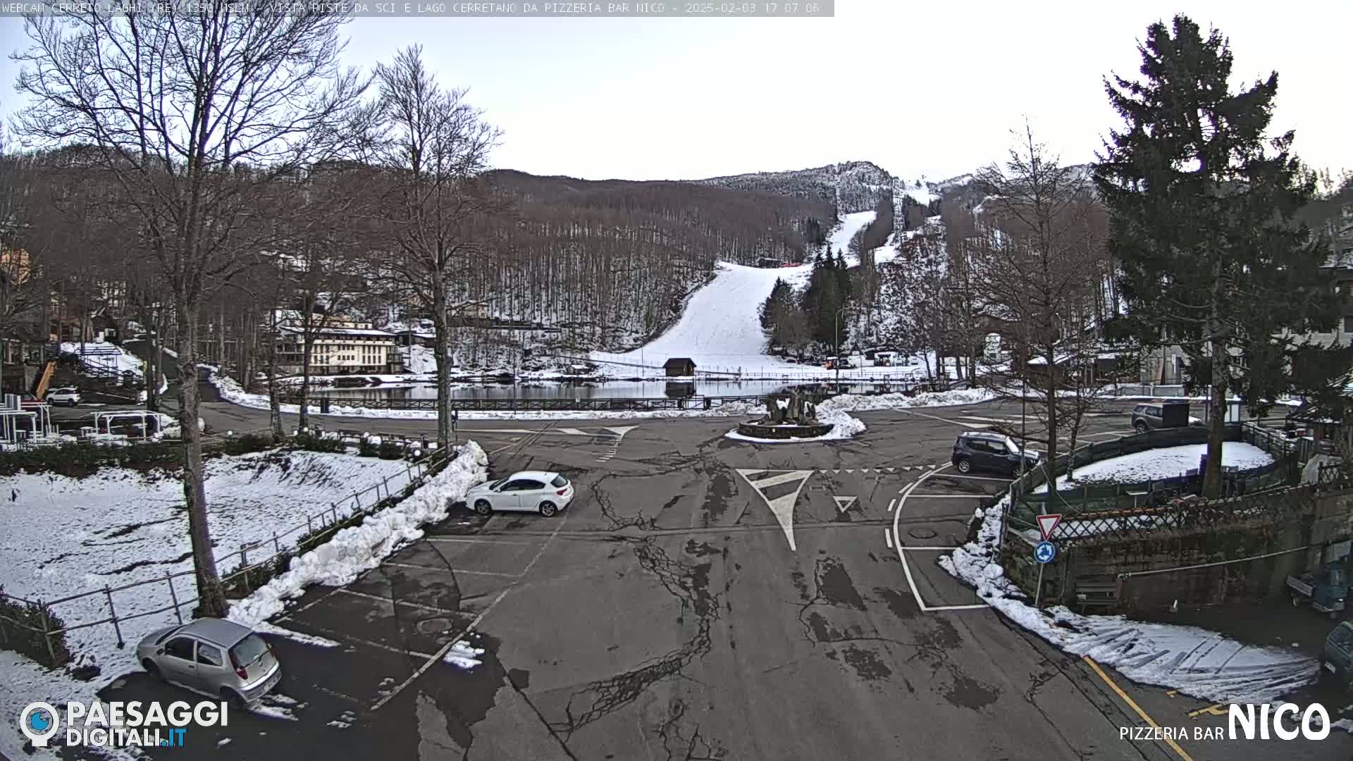 A snow-covered parking lot and roadway surround a small body of water, with a ski slope visible in the background under a cloudy sky.