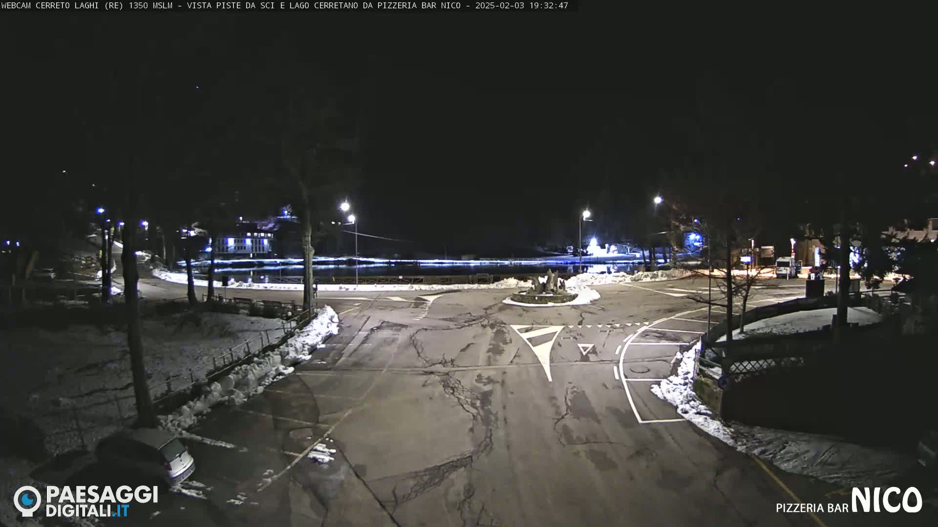 A nighttime view of a snow-covered parking lot and roadway surrounding a small body of water, illuminated by streetlights.