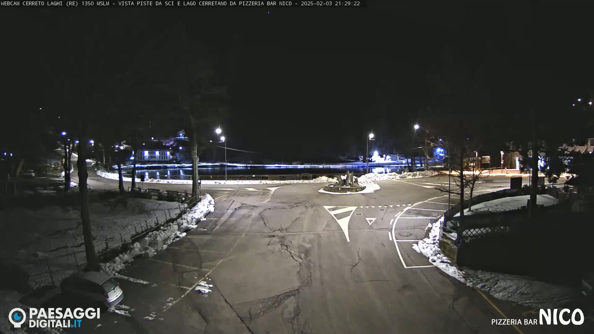 A nighttime view of a snow-dusted parking lot and roadway surrounding a small body of water, illuminated by streetlights.