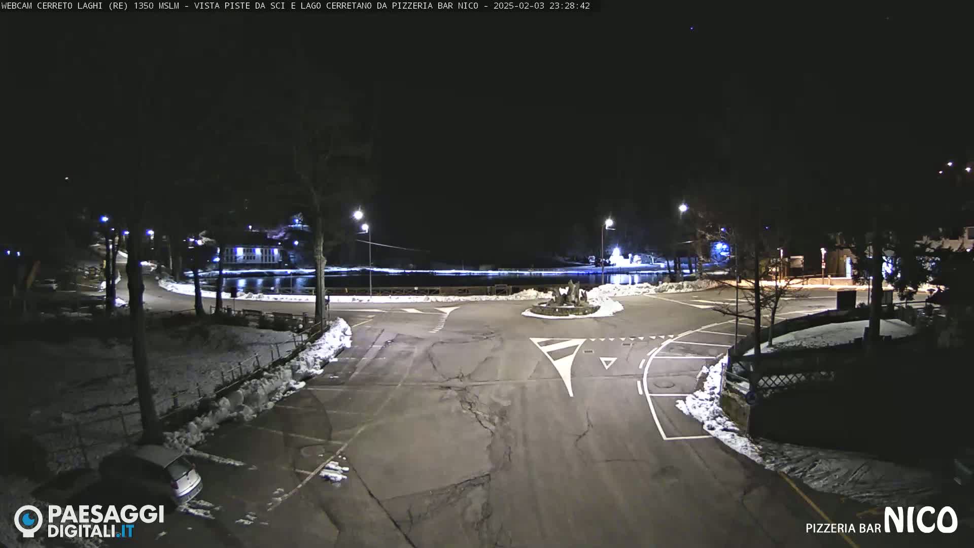 A nighttime view of a snow-covered parking lot and roadway surrounding a small lake, illuminated by streetlights.