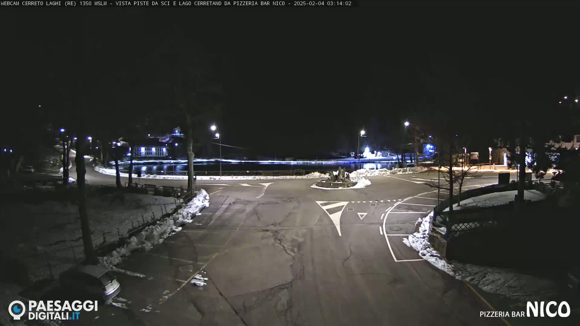 A nighttime view of a snow-covered parking lot and roadway surrounding a small lake, illuminated by streetlights.