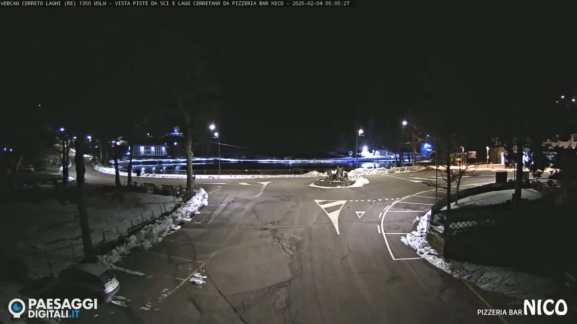 A nighttime view of a parking lot and roadway near a body of water, with some snow visible on the ground.