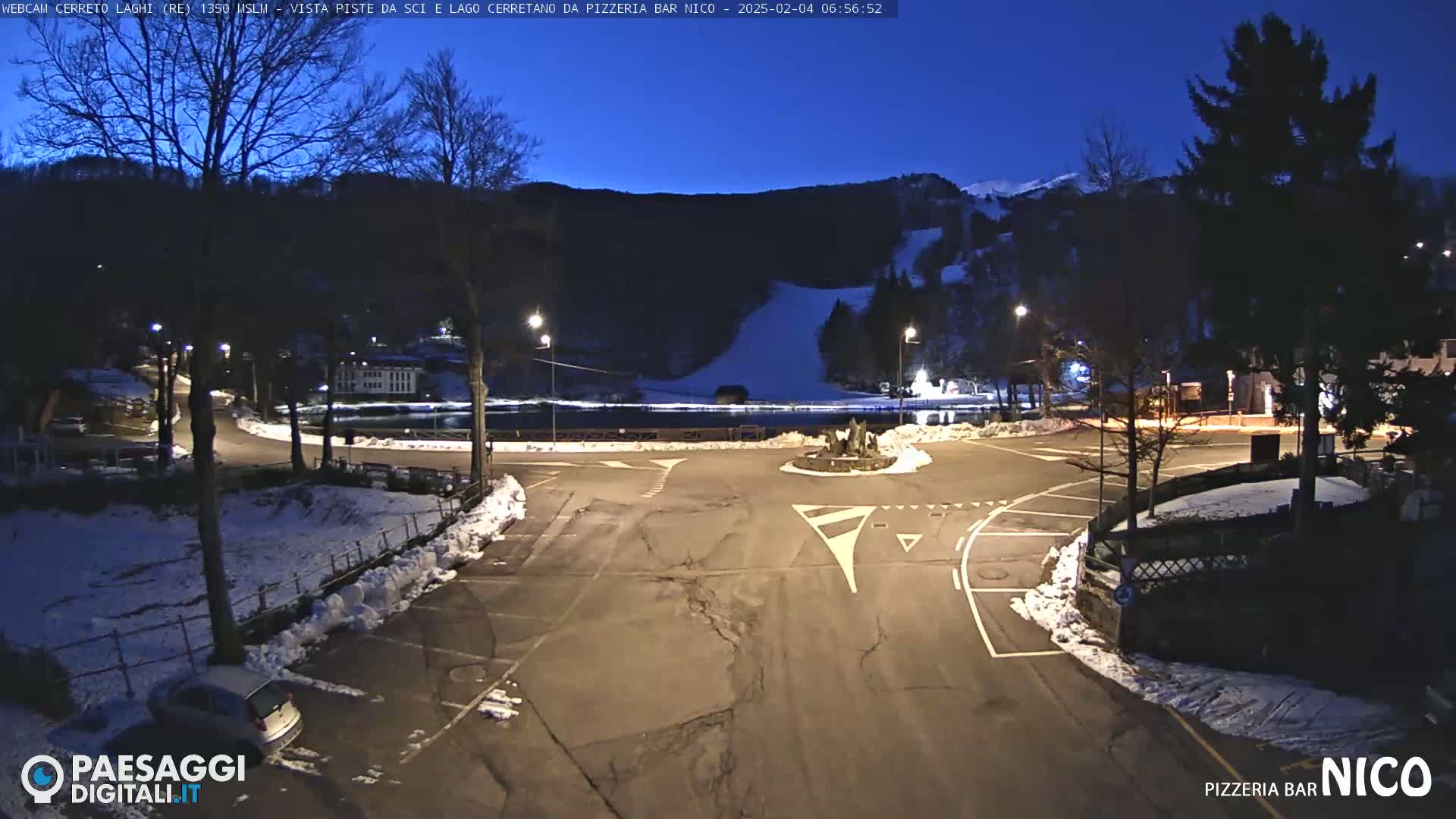 A snow-dusted parking lot and roundabout are illuminated at night, overlooking a dark ski slope and a body of water under a twilight sky.