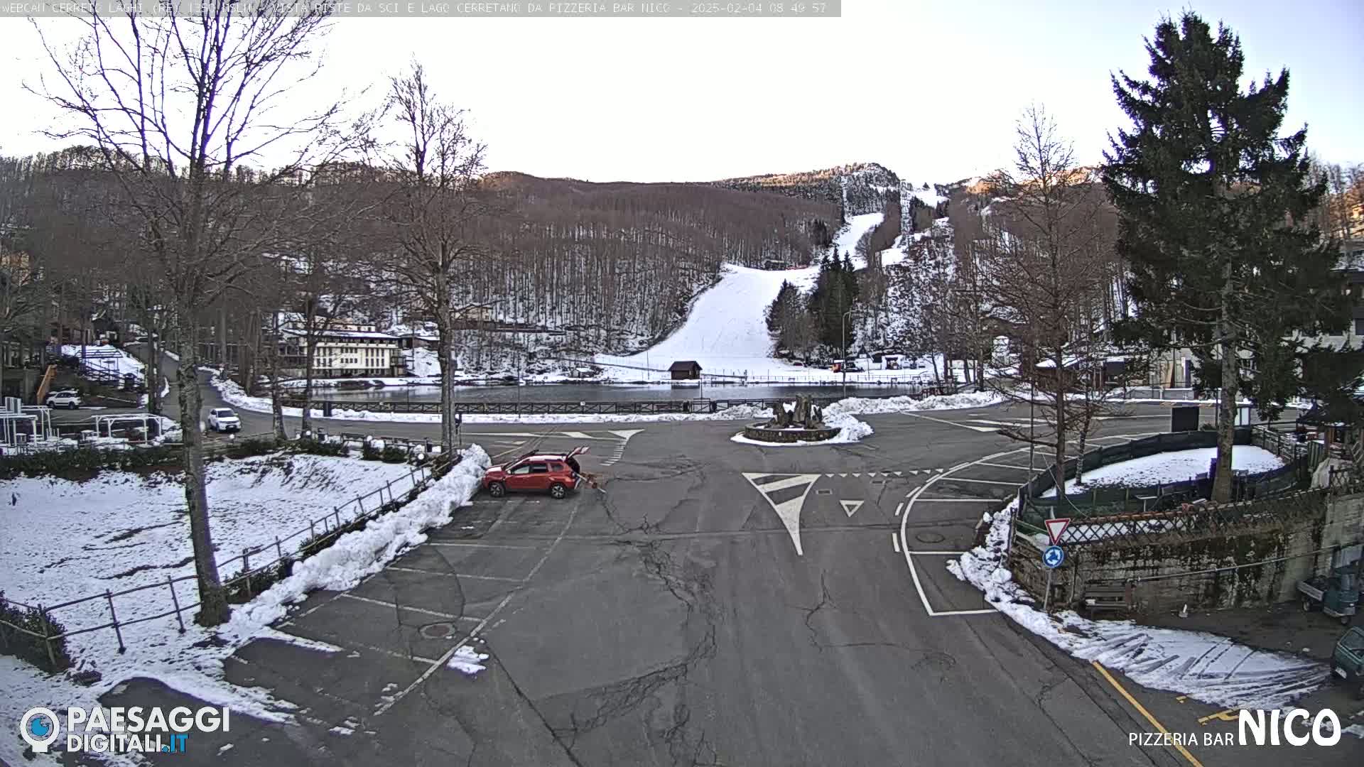 A mostly empty parking lot with a few cars, surrounded by snow-covered ground and bare trees, overlooks a snow-covered ski slope on a bright, clear day.