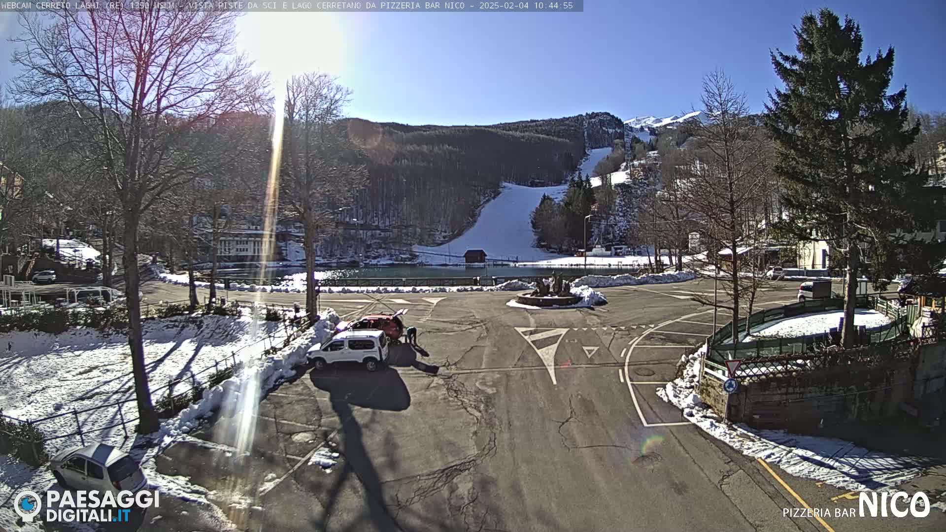 A sunny winter day reveals a parking lot with several cars, two people working on a red car, and a view of a snow-covered ski slope and surrounding mountains in the background.