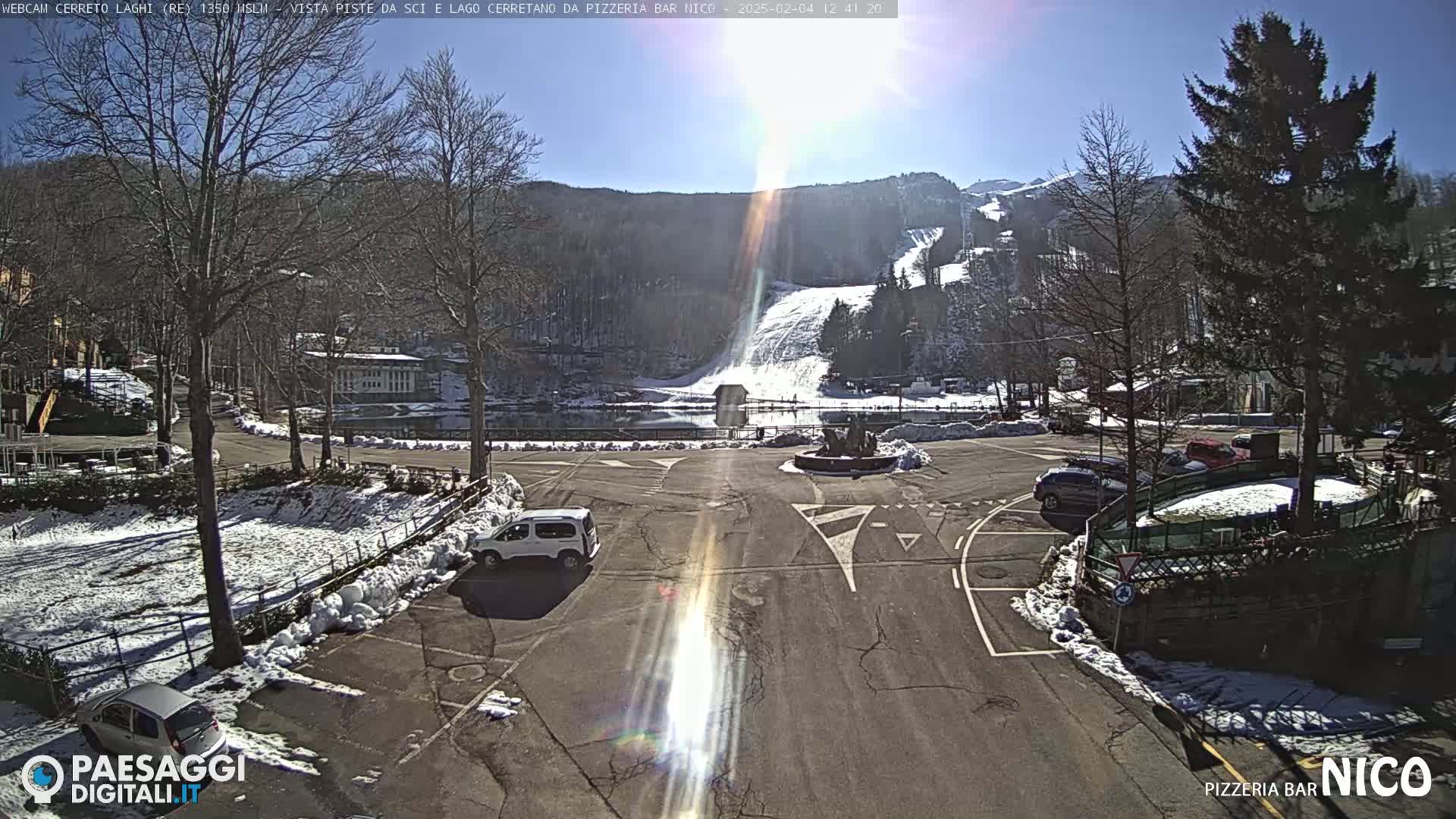 A sunny parking lot with some cars and snow on the ground, overlooking a ski slope and lake nestled in a snow-dusted mountain valley.
