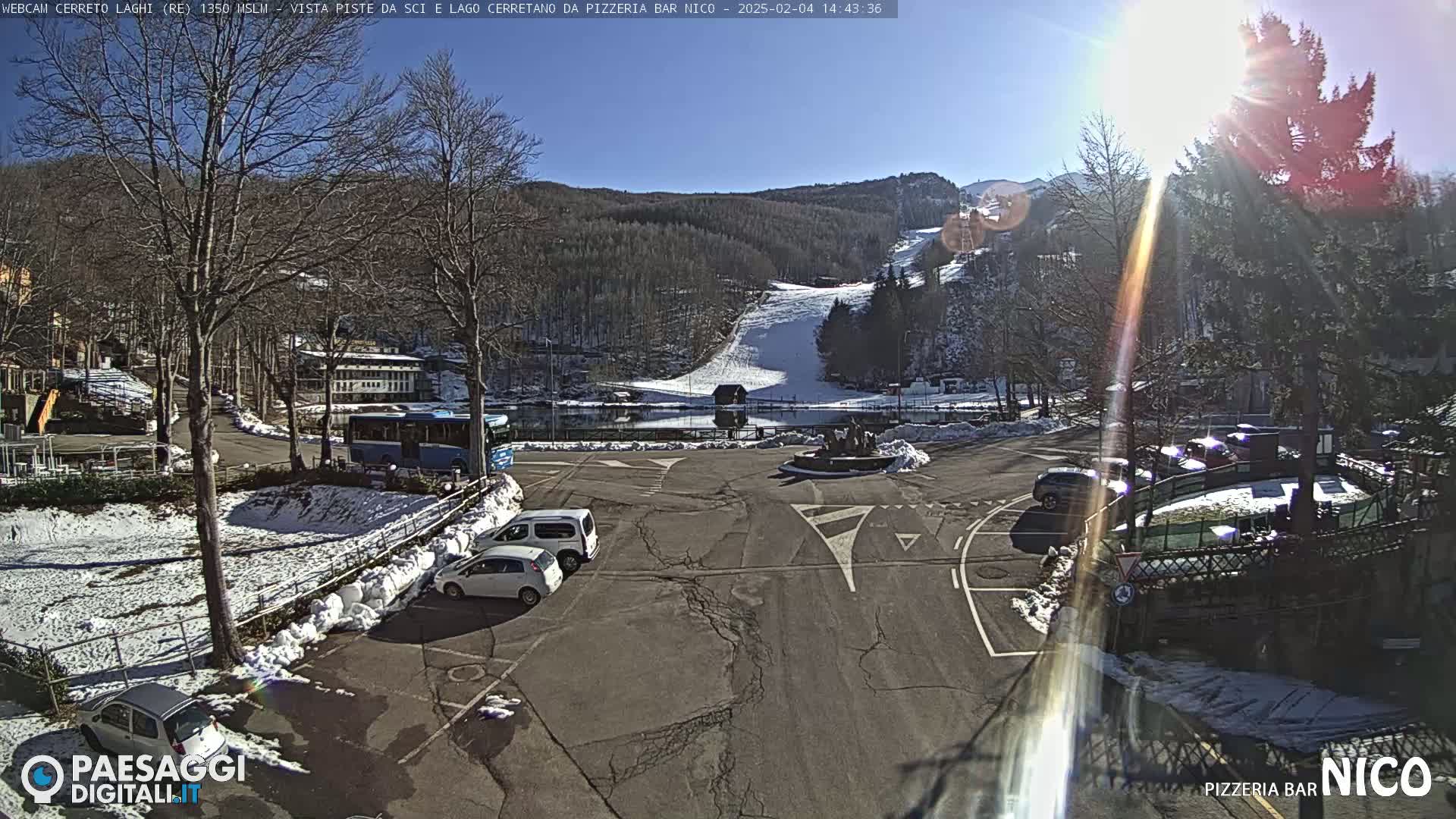 A sunny parking lot with several cars and a bus, surrounded by snow-covered ground and bare trees, overlooks a snowy ski slope and a lake in the background.